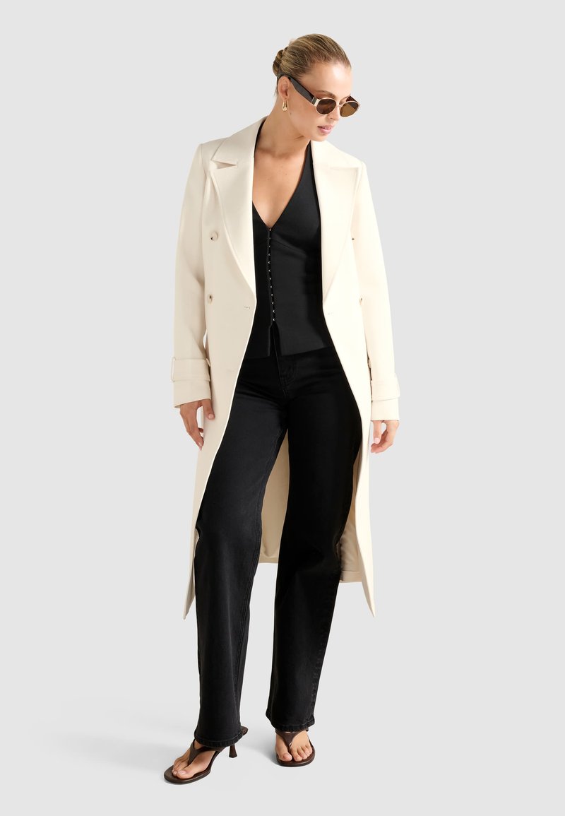 Frau in einem langen cremefarbenen Mantel, schwarzem V-Ausschnitt-Oberteil, schwarzen Jeans, braunen Absatzsandalen, Sonnenbrille und goldenen Creolen vor einem schlichten Hintergrund.