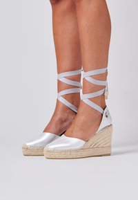 Wedge espadrille in satin argentato con suola in corda di juta e legami alla caviglia in raso. La parte superiore è liscia e presenta un design con punta rotonda.