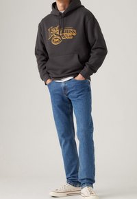 Tumma harmaa huppari, jossa on kultainen "Levi Strauss & Co." grafiikka, siniset denimit ja valkoiset lenkkarit punaisilla yksityiskohdilla. Rentoa istuvuutta ja rakennetta.