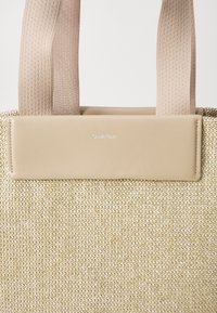 Bolsa de mano de tela beige con textura tejida, que presenta acentos de cuero crema suave y correas planas y resistentes. Logo de Calvin Klein grabado en la correa.