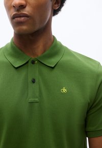 Nahaufnahme einer Person, die ein grünes Polohemd mit schwarzen Knöpfen und einem kleinen, gelben, aufgestickten Logo auf der Brust trägt.