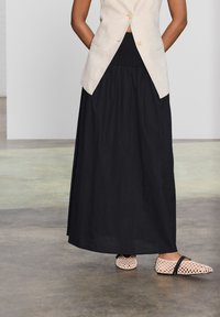 Jupe maxi noire avec une taille élastique, associée à un blazer en lin beige clair. Les chaussures sont de couleur claire, tissées avec une sangle sombre.