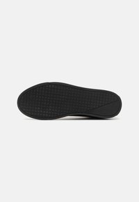 Semelle de chaussure noire avec un motif en grille couvrant la majeure partie du dessous et une zone lisse près du talon sur fond blanc.