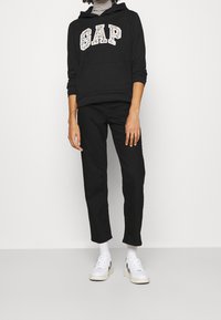 Sweat à capuche noir avec logo "GAP" imprimé animal, pantalon noir et baskets blanches avec accents noirs. Capuche et poche avant visibles.
