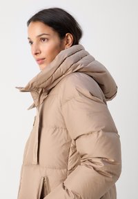 Beige puffjakker kõrge kraega, nähtava õmblusega, lukuga taskud ja läikiv viimistlus. Vasakul varrukal on logo.