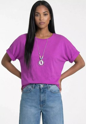 Kobieta z długimi czarnymi włosami stoi z rękami na biodrach, ubrana w fioletową koszulkę z krótkim rękawem, niebieskie dżinsy i kolorowy naszyjnik z zawieszką.