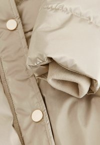 Cappotto puffer beige con una finitura lucida e testurizzata. Presenta un polsino a coste e grandi dettagli conbottoni dorati. Tessuto morbido e isolato.