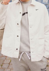 Person klädd i en vit knappad jacka över en grå Nike sweatshirt och grå sweatpants med vit dragsko, stående på kullersten.