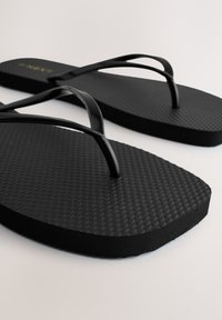Tongs en caoutchouc noir texturé avec des fines lanières brillantes, photographiées sur une surface blanche.