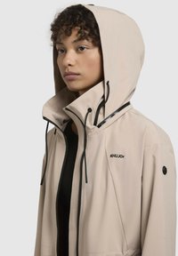Giacca leggera beige resistente alle intemperie con cappuccio, caratterizzata da dettagli neri, cerniere e un logo sul petto.