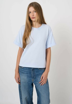 Młoda kobieta z długimi blond włosami, ubrana w jasnoniebieską koszulę z krótkim rękawem i luźne niebieskie dżinsy, na jednolitym białym tle.