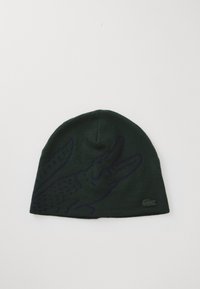 Bonnet tricoté vert avec un graphique de crocodile violet et un petit logo Lacoste sur le côté. Texture douce, coupe ajustée, sommet arrondi.