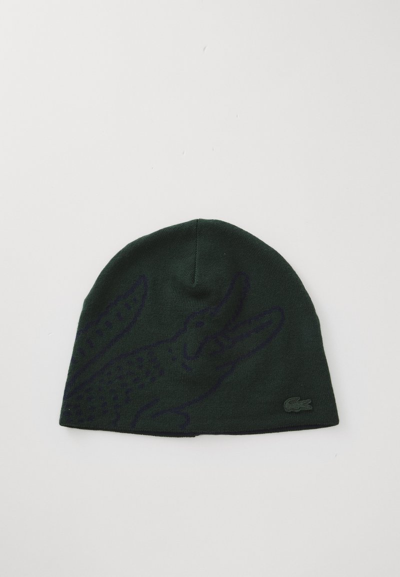 Bonnet tricoté vert avec un graphique de crocodile violet et un petit logo Lacoste sur le côté. Texture douce, coupe ajustée, sommet arrondi.
