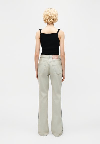 Άτομο με κοντά κυματιστά ξανθά μαλλιά φοράει μαύρο αμάνικο τοπ και ανοιχτό γκρι τζιν παντελόνι με ψηλή μέση και φαρδιά πόδια, στρέφεται με την πλάτη προς τα εμπρός.