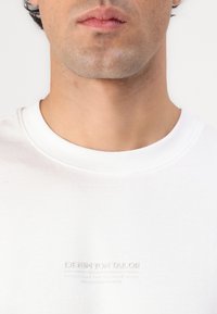 Hvid bomulds t-shirt med ribbet rund halsudskæring og prægede logo detaljer på brystet, der viser en glat tekstur og minimalistisk design.