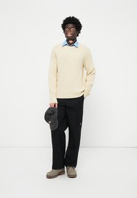 Cream ribbed sweater, light blue collared shirt, black cargo pants, grey cap in hand, and light brown boots, standing against a white background.