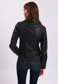 Giubbotto in pelle nera con un design aderente, dettagli trapuntati sulle spalle e colletto classico. Indossato sopra un paio di jeans in denim blu scuro.