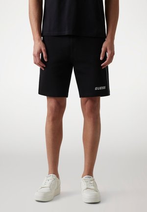 Juodos šortai su elastingu juosmeniu, šoninėmis baltomis juostelėmis ir sidabriniu "GUESS" logotipu kairėje kojoje. Pora su baltomis sportbačiais.