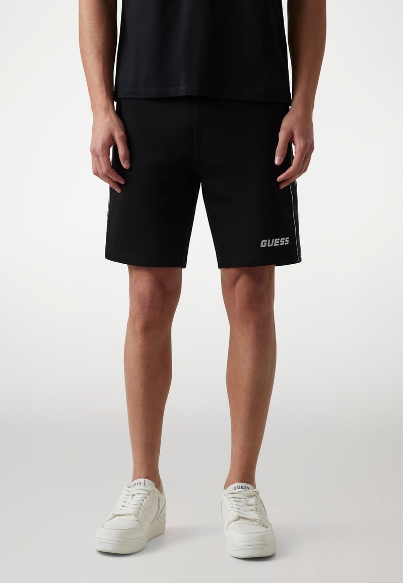 Czarne szorty z elastycznym paskiem w talii, białe paski po bokach i srebrne logo "GUESS" na lewej nodze. W połączeniu z białymi sneakersami.