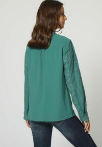 Blusa de color azul verdoso con mangas largas bordadas que presentan patrones florales. Tejido ligero con corte holgado y dobladillo redondeado.