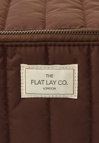 Brązowa pikowana tkanina z widocznym zamkiem błyskawicznym, z białą metką, na której czarnym tekstem napisano "THE FLAT LAY CO. LONDON".