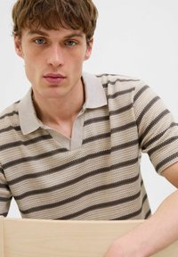 Polo cu dungi, în nuanțe de bej și maro, dintr-un material texturat, cu mâneci scurte, guler în formă de V, pe un fundal cu textură de lemn vizibilă.