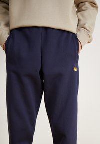 Marineblaue Jogginghosen aus weichem Material mit Seitentaschen und einem kleinen gelben Logo am linken Oberschenkel. Elastischer Bund für mehr Komfort.