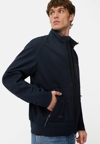 Ung mand iført en navy blå hættetrøje med lynlås og hånden i lommen, kigger mod sin højre side, mod en ensfarvet hvid baggrund.