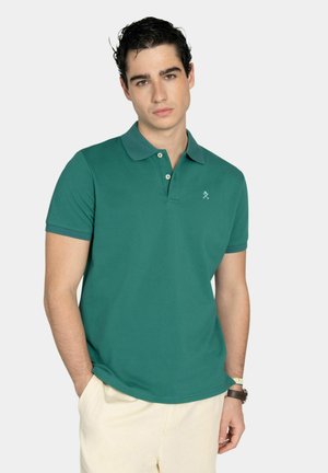 Joven con cabello oscuro, vestido con una camisa polo verde y pantalones beige, de pie con las manos en los bolsillos frente a un fondo liso.