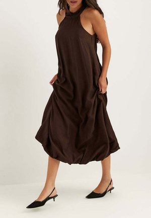 Kvinde iført en ærmeløs mørkebrun midi-kjole med høj hals og sorte lavhælede slingback-sko, der går mod en ensfarvet baggrund.