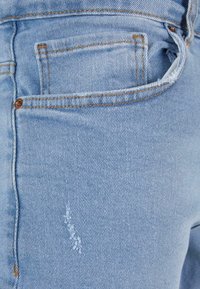 Jean denim bleu clair avec poches cousues et un effet usé. Coupe standard avec couture visible et texture subtile.