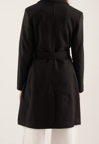 Černý vlněný kabát s páskem v pase, dlouhými rukávy a zadním rozparkem. Tkanina má měkkou texturu a strukturovaný tvar.
