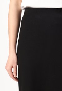 Svart stretch blyantskjørt med høy midje, med en glatt tekstur og elegant design. Hånd plassert ved skjørtets side.