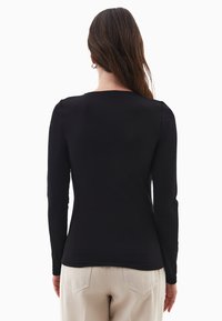 Donna con lunghi capelli castani, che indossa una semplice maglietta nera a maniche lunghe e pantaloni beige, in piedi di spalle su uno sfondo bianco.