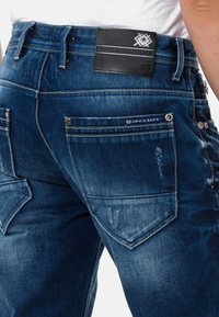 Dunkelblaue Denim-Jeans mit einer abgenutzten Textur, mit einem ledernen Markenaufnäher, mehreren Taschen und metallischen Akzenten auf der Rückseite.