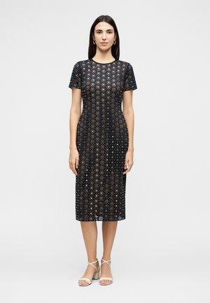 Γυναίκα που στέκεται φορώντας ένα ναυτικό μπλε φόρεμα μέχρι το γόνατο με κοντά μανίκια και με σχέδιο ανθών με κοψίματα και μπεζ σανδάλια με λουράκια και τακούνι τύπου block.