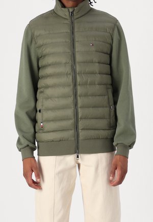 Homme portant une veste matelassée zippée vert olive à manches longues et un pantalon beige, debout devant un fond uni.