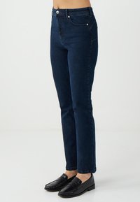 Jeans slim fit blu scuro indossati con mocassini in pelle nera, mostrati dalla vita ai piedi contro uno sfondo chiaro e semplice.