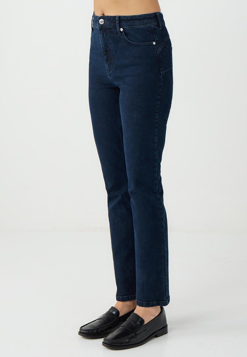 Jeans slim fit blu scuro indossati con mocassini in pelle nera, mostrati dalla vita ai piedi contro uno sfondo chiaro e semplice.