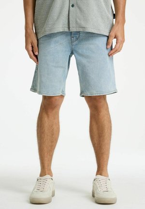 Man wearing light blue denim shorts, a gray patterned shirt, and white sneakers, standing against a plain white background.