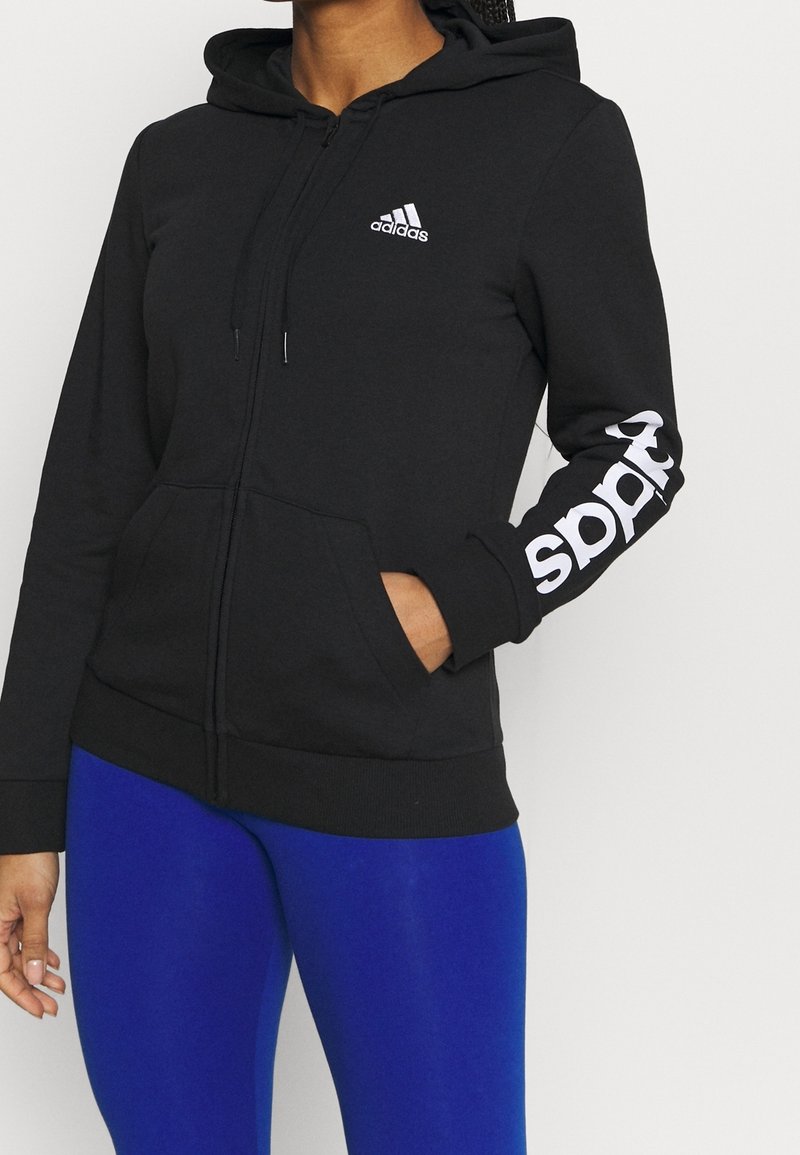Sweat à capuche zippé Adidas noir avec logo blanc sur la poitrine et la manche, porté avec des leggings bleu vif, mains dans les poches avant.