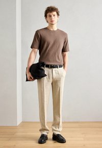 Brown short-sleeve t-shirt, beige linen trousers, black belt, dark shoes, holding a black bag; minimalistic design against a light background.