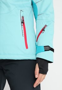 Veste bleu clair avec poches zippées rouges, sangle de poignet ajustable, portée sur un pantalon noir avec trou pour le pouce au bas de la manche.