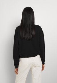 Persona con lunghi capelli neri lisci, che indossa una felpa nera e pantaloni bianchi, in piedi di fronte a una parete grigia chiaro uniforme.