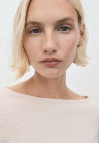 Primo piano di una persona dalla pelle chiara con capelli biondi, che indossa un maglione lavorato a maglia di un beige chiaro, con un ampio scollo e una texture morbida.