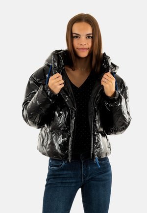 Giacca a vento nera con finitura lucida, design cropped, cappuccio regolabile e dettagli blu. Indossata con una maglietta a pois scuri e jeans.