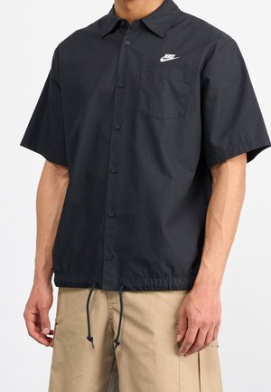 Homme portant une chemise noire à manches courtes avec boutons et logo Nike, ainsi qu'un short cargo beige, debout devant un fond blanc.