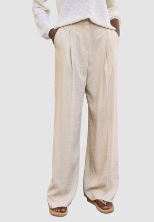 Pantalons large beige clair en tissu texturé, avec des plis à l'avant et des poches latérales. La ceinture présente un détail de fermeture discret.