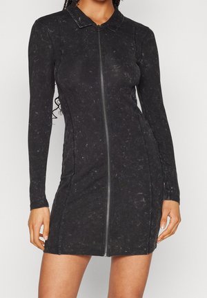 Femme portant une robe mini noire cintrée à manches longues avec une fermeture éclair frontale et un col, debout devant un fond clair uni.