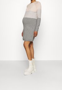 Gravid person som står, iklädd en långärmad, flerfärgad stickad klänning i nyanser av grått och beige, tillsammans med vita stövletter.